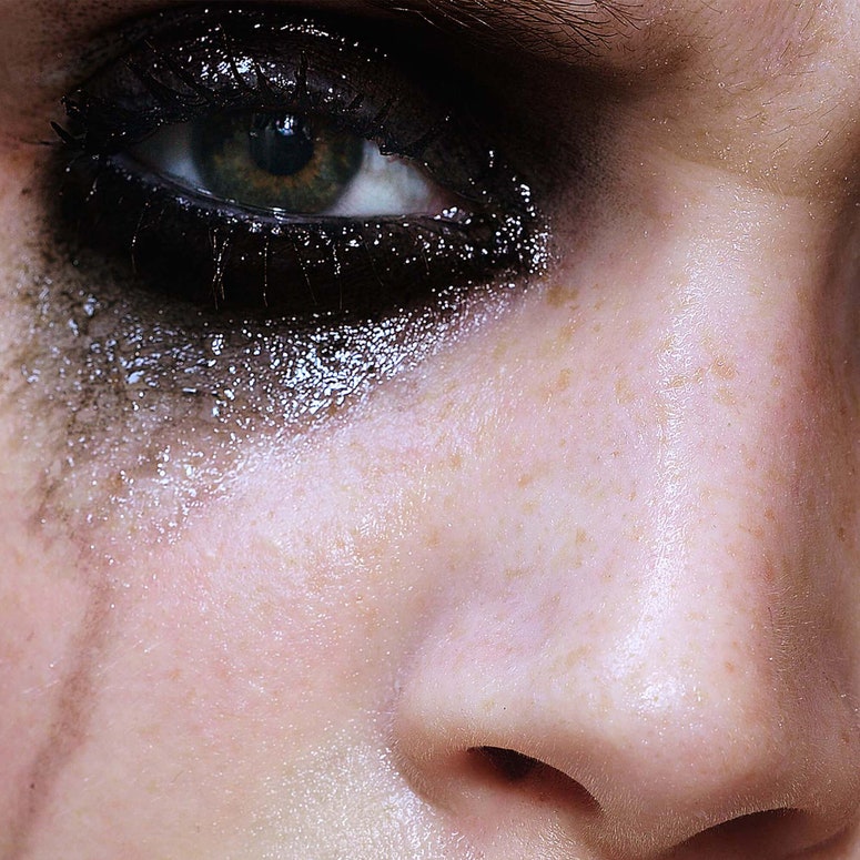 A close-up image of a white woman’s face. She is wearing dark, smudged eyeshadow, with silver glitter running down her cheek in tear stains. 