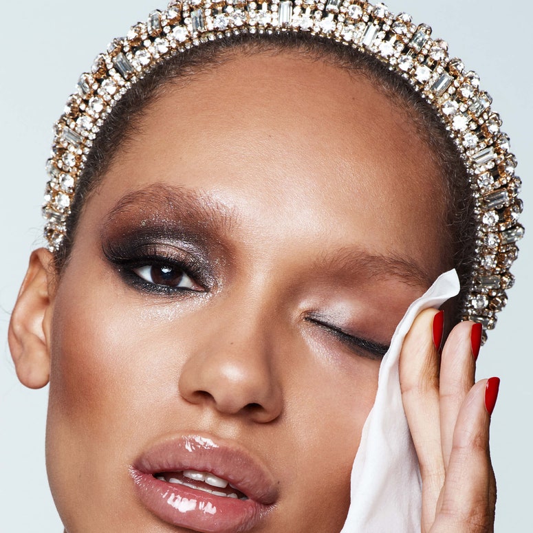 A mixed race woman with glamorous makeup and a bedazzled headband wipes makeup off her left eyelid. 